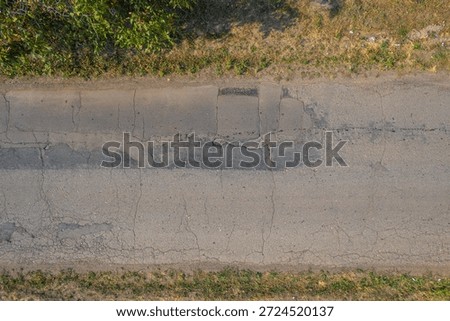 A top-down view of an old worn asphalt road featuring cracks, patched sections and dry grass along the edges, illustrating aging infrastructure and rural road conditions.