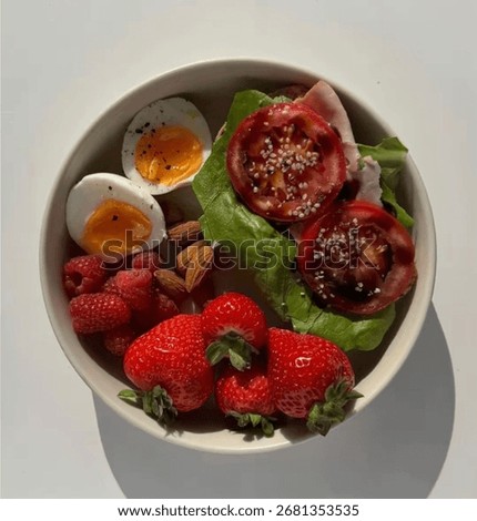 Top-down view on a white background of a fresh, colorful plate with tomatoes, boiled egg, leafy salad, strawberries, raspberries, and almonds—nutritious, vibrant, and full of flavors.