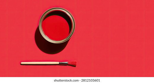 A top-down view of an open can filled with vivid red paint, placed next to a clean paintbrush on a solid red surface. The scene is illuminated by strong light, casting a distinct shadow.