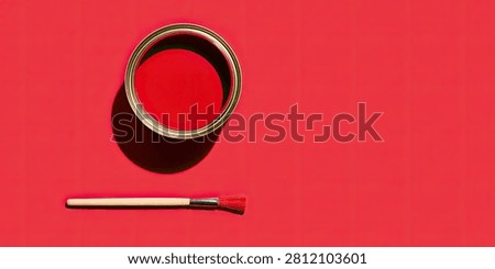 A top-down view of an open can filled with vivid red paint, placed next to a clean paintbrush on a solid red surface. The scene is illuminated by strong light, casting a distinct shadow.