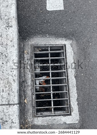 A top-down view of a rectangular metal storm drain grate on an asphalt surface next to a concrete curb. Debris is visible inside, illustrating urban infrastructure and a gritty, industrial concept.