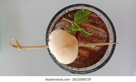 Top-down view of a refreshing iced lychee tea served in a clear glass. The drink is garnished with a skewer holding a whole peeled lychee fruit and a fresh mint leaf