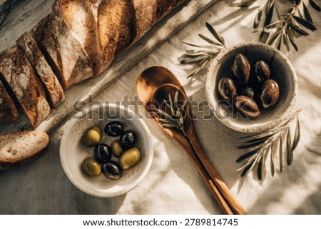 A top-down view of a rustic sourdough loaf, sliced and served with bowls of black and green olives, garnished with fresh rosemary sprigs and wooden spoons on a textured linen cloth.