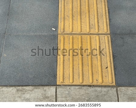 A top-down view of a section of a city sidewalk. The image features a bright yellow tactile pavement with raised lines, designed to assist visually impaired people. The textured surface contrasts
