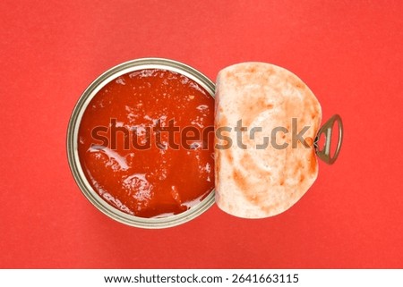 A top-down view shows an open can of chunky red tomato sauce with its lid peeled back, set against a vibrant red background