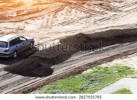 Top-down view of silver station wagon car parked next to multiple dark piles of asphalt chips used for road repair on sandy ground. Tire tracks crisscross dirt, sun flare illuminates scene.
