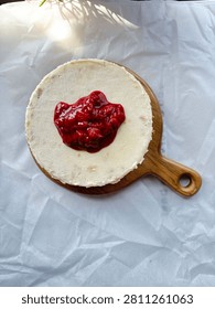 Top-down view of a single serving strawberry cheesecake with red berry jam topping on a round wooden board, placed on a white cloth. A delicious, creamy homemade Indonesian dessert perfect for food, bakery, recipe, and culinary stock photography.