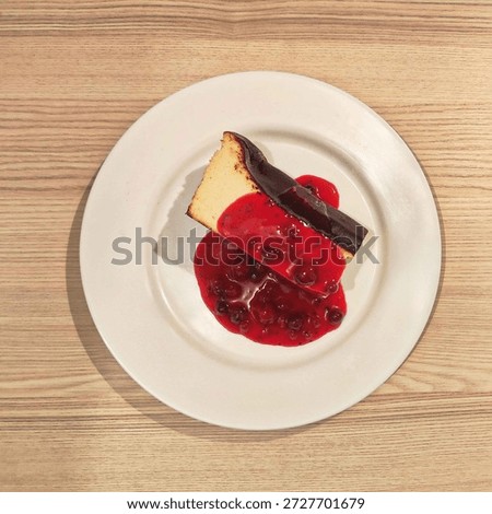 Top-down view of a slice of Basque burnt cheesecake covered in red berry coulis. Minimalist dessert photography on a white plate and wooden table.