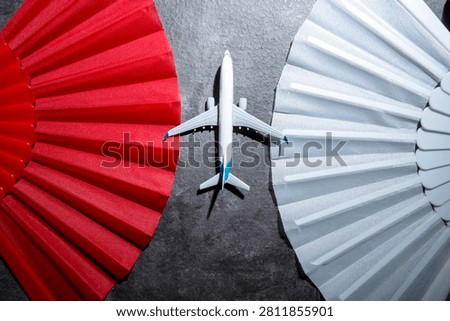 A top-down view of a small model airplane placed in the center between a vibrant red folding fan and a patterned white fan on a textured gray background, symbolizing travel and choices
