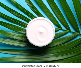A top-down view of a small, round jar of pale pink cream. The jar is centered on a large, vibrant green palm frond, which is laid out against a bright blue background. The lighting is soft and even, highlighting the textures of the frond and the smooth surface of the cream.