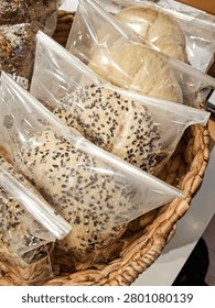 A top-down view of soft, baked bread rolls and bagels individually wrapped in clear plastic bags, arranged inside a rustic woven basket