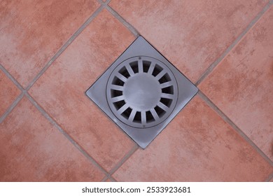 A top-down view of a square floor drain with a circular grill on reddish-brown tiled flooring. The drain design is geometric and functional, blending with the clean, tiled surface