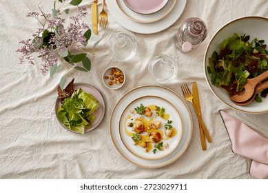 Top-down view of a stylish dining setup featuring a floral arrangement, gold cutlery, water glasses, pink accessories, and plated dishes including seared scallops and fresh salads on elegant white and pink tableware