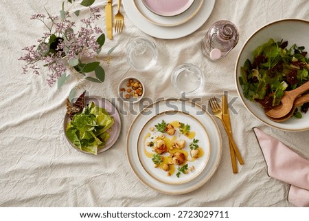 Top-down view of a stylish dining setup featuring a floral arrangement, gold cutlery, water glasses, pink accessories, and plated dishes including seared scallops and fresh salads on elegant white and pink tableware