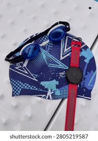 Top-down view of swimming essentials including blue swimming goggles, a patterned swim cap, and a sports smartwatch arranged on a poolside starting block with water droplets. Ideal for themes of swimming, fitness, aquatic training, healthy lifestyle, sports equipment, and exercise.