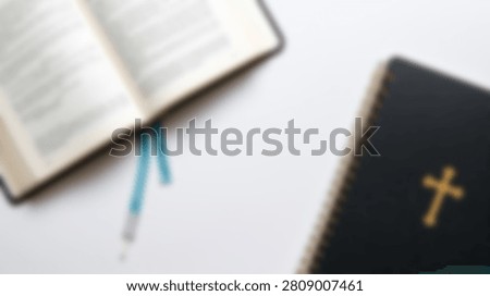 A top-down view of two Bibles on a clean white surface. One Bible is open, revealing its pages and a blue bookmark, while the other is closed, displaying a golden cross on its dark cover.
