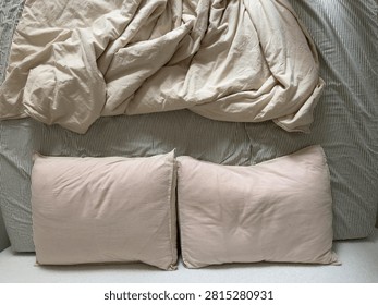 Top-down view of two cream pillows and a rumpled cream duvet on a striped green and cream sheet. The relaxed bedding evokes a cozy, slept-in bed.