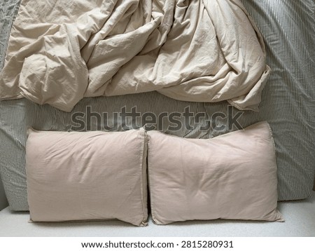 Top-down view of two cream pillows and a rumpled cream duvet on a striped green and cream sheet. The relaxed bedding evokes a cozy, slept-in bed.