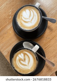 Top-down view of two flat whites with latte art in ceramic cups on black saucers, served on a wooden table in a cozy café, perfect for a relaxed coffee moment for two.