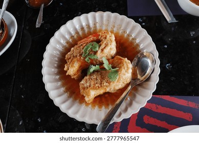 A top-down view of two large cooked prawns covered in savory brown garlic sauce and garnished with fresh green cilantro, served in a fluted white bowl with a metal spoon on a dark restaurant table.