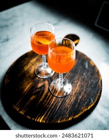 Top-down view of two Paper Plane cocktails in elegant stemmed glasses on a rustic wooden board. Bright orange hue + lemon twist garnish, ideal for themes of mixology, craft cocktails, and bar settings