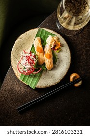 Top-down view of two pieces of flame-seared salmon nigiri topped with orange tobiko roe, served on a speckled ceramic plate alongside chopsticks and a glass of sparkling wine.