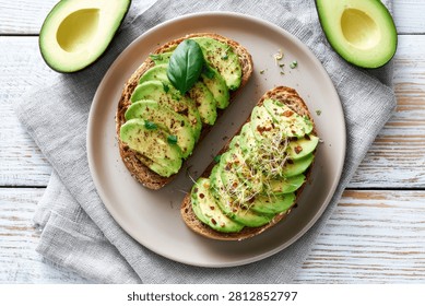A top-down view of two slices of avocado toast, artfully arranged with thin avocado slices, fresh herbs, and seasoning, served on a rustic plate.