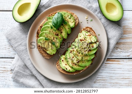 A top-down view of two slices of avocado toast, artfully arranged with thin avocado slices, fresh herbs, and seasoning, served on a rustic plate.