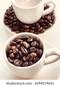 Top-down view of two white coffee cups: one filled with coffee and milk, and the other with whole roasted coffee beans, set on white saucers.