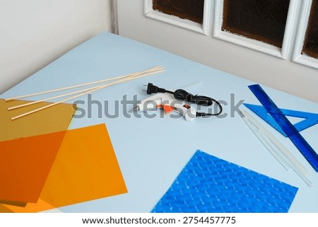 Top-down view of various DIY craft supplies ready for a project. A hot glue gun, rulers, vibrant orange blue plastic sheets sit on a light blue desk. Clean, bright lighting. Creative hobby concept.