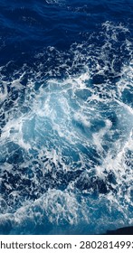 top-down view of vibrant deep blue ocean water. Dynamic white sea foam and intricate bubble patterns swirl across the surface, created by the churning motion of waves or a passing boat. The contrasting colors deliver a refreshing and energetic coastal atmosphere