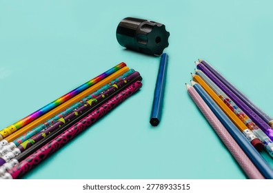 A top-down view of vibrant, patterned pencils arranged symmetrically around a modern black manual sharpener on a clean turquoise surface.