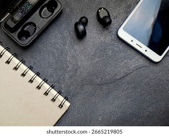 A top-down view of a workspace featuring wireless earbuds, a smartphone, a charging case, and a notebook on a dark textured surface. The image suggests technology, productivity, and modern lifestyle w