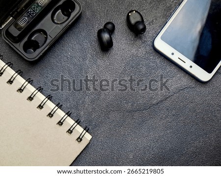 A top-down view of a workspace featuring wireless earbuds, a smartphone, a charging case, and a notebook on a dark textured surface. The image suggests technology, productivity, and modern lifestyle w