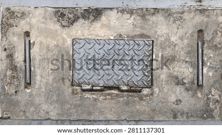 A top-down weathered view of a heavy concrete slab serving as a ground access panel, featuring a central rectangular metal diamond plate hatch door with hinges