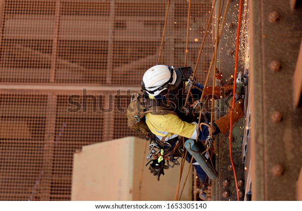 Tope view of rope access technician welder services\
wearing white helmet head fall protection PPE, face shield\
abseiling working at height using power grinder grinding\
preparation prior to welding  \
