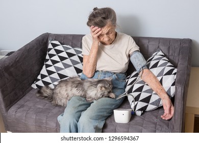 The Topic Is Very Old Person And Health Problems. A Senior Caucasian Woman, 90 Years Old, With Wrinkles And Gray Hair, Sits Home On Sofa With Pet Cat And Uses Tonometer. High Blood Pressure Headache.