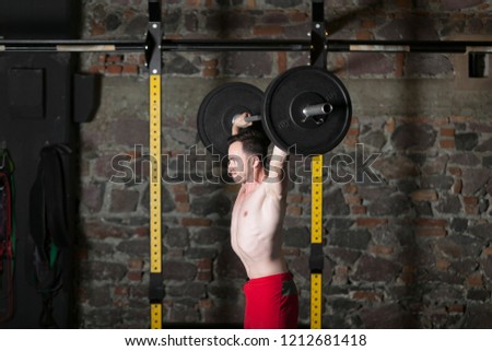 Topless male athlete practicing olympic lifts at gym with brick wall background.