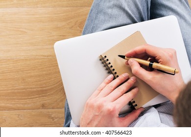 Topview of man writing in aged notepad placed on closed laptop. Wooden background