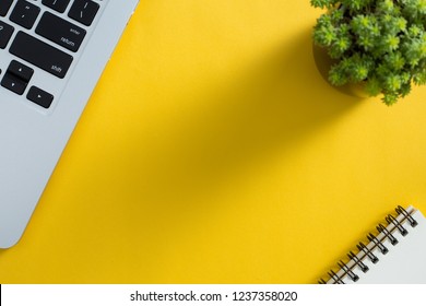 Topview Of Yellow Office Desktop With Laptop Keyboard, Green Plant And Notepad.