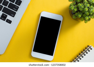 Topview Of Yellow Office Desktop With White Smartphone, Laptop Keyboard, Green Plant And Notepad.