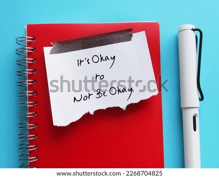 Torn note paper stick on red notebook with text written IT'S OKAY TO NOT BE OKAY, means feelings and emotions expressing are valid no matter what they are, it is normal to say you are not okay