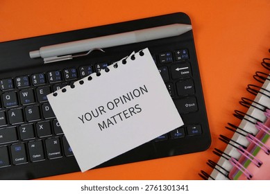 A torn note reading 'Your Opinion Matters' placed on a computer keyboard with a pen and spiral notebooks on an orange desk, conveying feedback, survey participation and workplace communication.