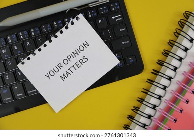 A torn note reading 'Your Opinion Matters' placed on a computer keyboard with a pen and spiral notebooks on an yellow desk, conveying feedback, survey participation and workplace communication.