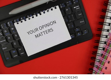 A torn note reading 'Your Opinion Matters' placed on a computer keyboard with a pen and spiral notebooks on an red desk, conveying feedback, survey participation and workplace communication.
