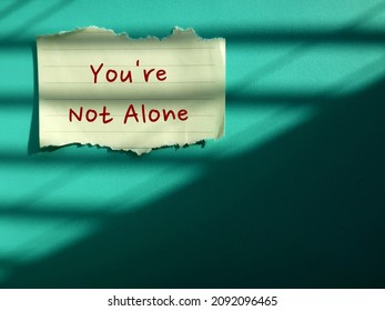 Torn White Stick Note On Blue Background Written YOU'RE NOT ALONE - Concept Of Positive Self Talk Or Affirmation, Encouragement Message To Support Other People When Life Seems Difficult And Hopeless