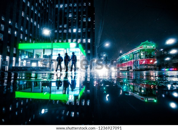TORONTO CITY NIGHT SCENE - Silhouetted figures\
walking past bright neon light building with Toronto streetcar\
vehicle driving by. Urban downtown scene, reflections in puddle.\
Toronto, Ontario,\
Canada
