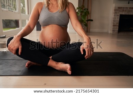 Torso of Pregnant Caucasian female sitting cross legged on yoga mat while holding her knees. Healthy lifestye in modern home.