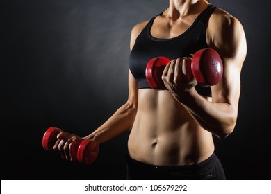 Torso of a young fit woman lifting dumbbells on dark background