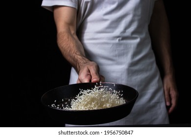 Toss The Rice In The Skillet. The Cook Is Throwing Rice On A Black Background. Cooking. Photo Of Food On A Dark Background